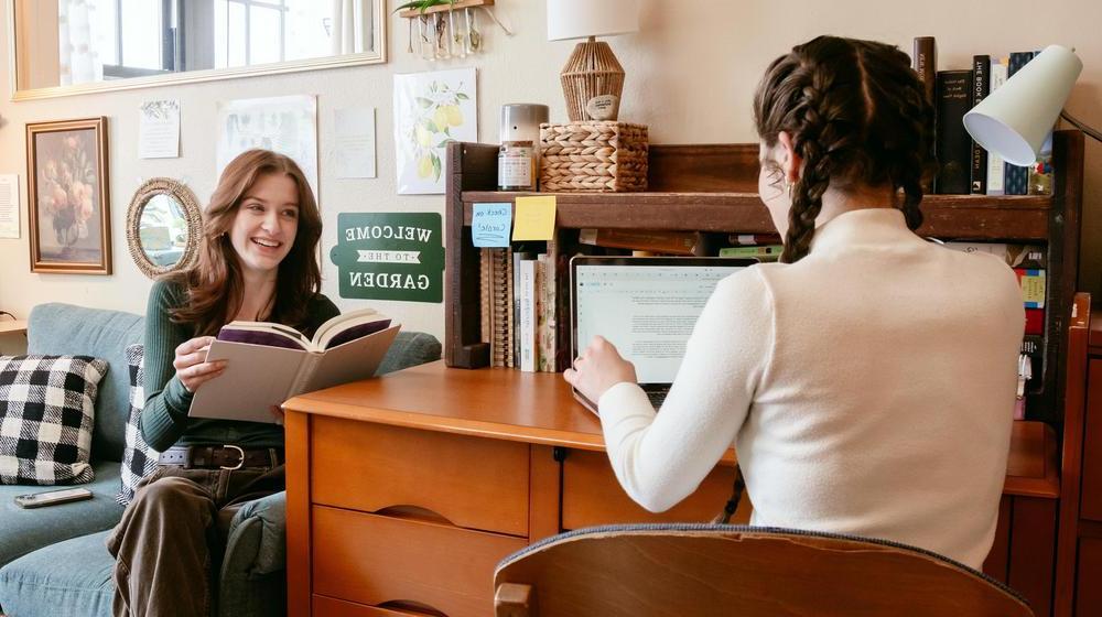 Two students in a dorm room, one working on a laptop and the other reading a book, chatting together.